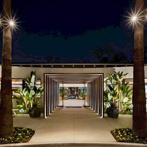 A modern entrance at night, featuring a pathway with illuminated structural frames, flanked by two palm trees and greenery, leading to a building.