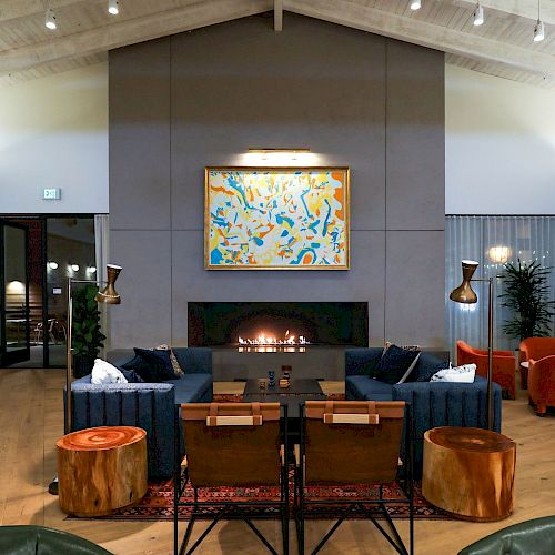 A modern living room with high ceilings, a central fireplace, a colorful painting, and stylish furniture including blue sofas and wooden tables.