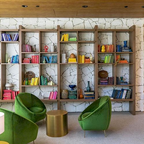 A modern living room with colorful books and decor on shelves, green chairs, a small table, and a lamp. The wall has a stone texture.