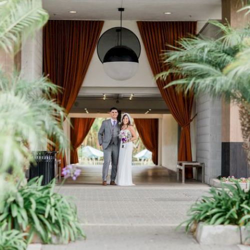 A couple stands together in a well-decorated hallway with curtains and plants on either side, possibly at an event or wedding.
