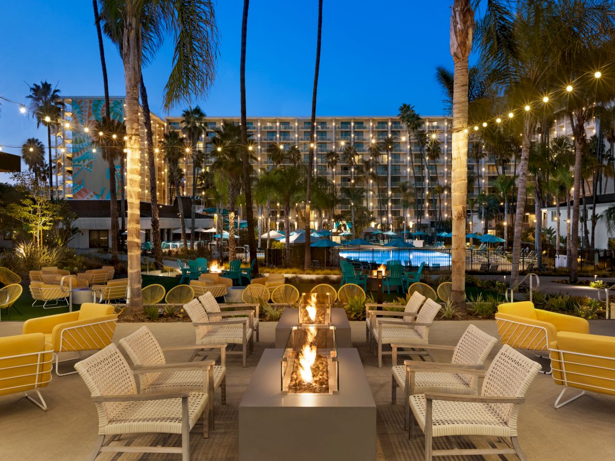 Palm-lined hotel patio at dusk with a fire pit, string lights, teal umbrellas, and a lit pool beside guest rooms.