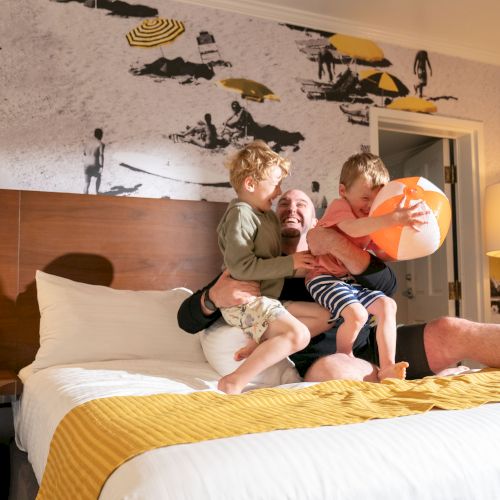 A person and two children laughing on a bed, one holding a beach ball, in a brightly lit room with beach-themed wall art.