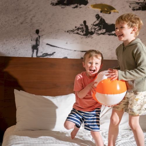 Two smiling children are jumping on a bed, holding an orange and white beach ball. Decor includes a black-and-white mural and a nightstand flower.