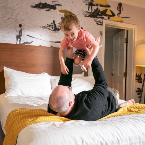 A person lies on a bed, lifting a happy child into the air in a warmly lit room decorated with a beach-themed mural.