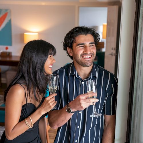 Two people are standing near a window, smiling and holding glasses of champagne in a well-lit room with modern decor.