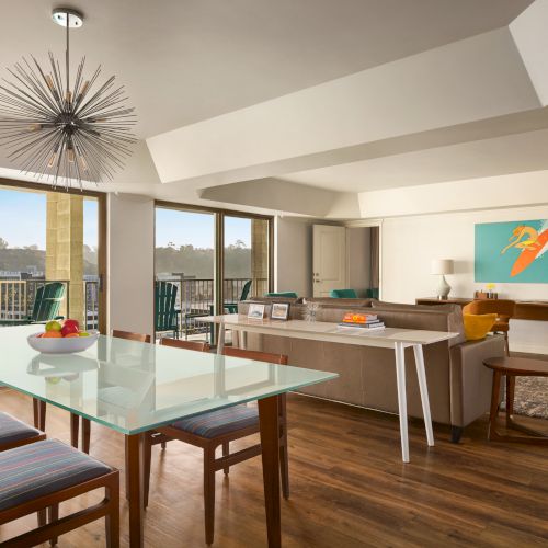 A modern living room with a glass table, colorful chairs, a unique light fixture, and artwork on the wall, opening to a balcony.
