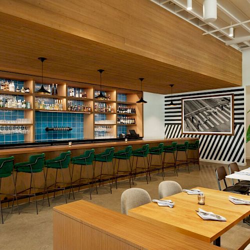 The image depicts a modern bar and dining area with green barstools, wooden tables, and a wall with shelves filled with various bottles.