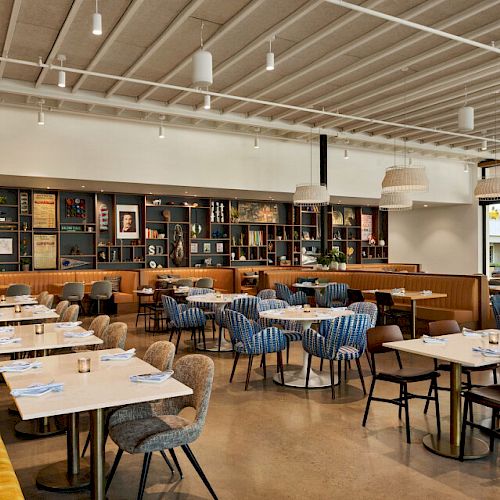 The image shows a modern, well-lit restaurant with a mix of yellow booth seating, blue-striped chairs, and bookshelves lining the back wall.