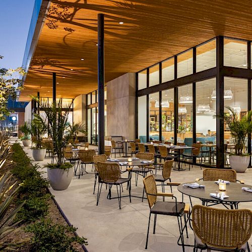 An outdoor dining area with plenty of tables and chairs, surrounded by plants, adjacent to a modern glass-walled building with indoor seating.
