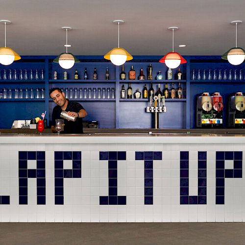 A bartender prepares a drink behind a white counter with "LAP IT UP" written on it, with various bottles and drink dispensers on the shelves.