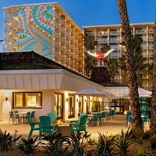 The image shows a resort with outdoor seating, a mural on a tall building, and palm trees surrounding the area at dusk, lit with warm lights.