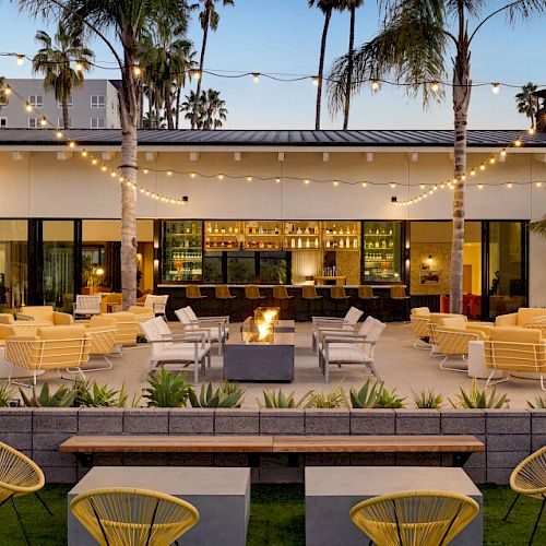 Outdoor patio with yellow chairs, palm trees, string lights, fire pit, and a bar in the background, creating a relaxed, modern ambiance.