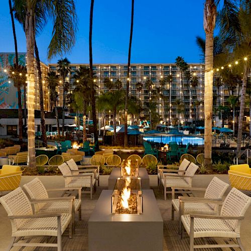 An outdoor lounge area with a fire pit, surrounded by seating, palm trees, and string lights, facing a pool with a hotel in the background illuminated at night.