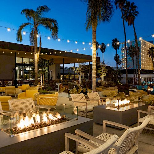 An outdoor seating area with fire pits, string lights, palm trees, and buildings in the background at twilight.