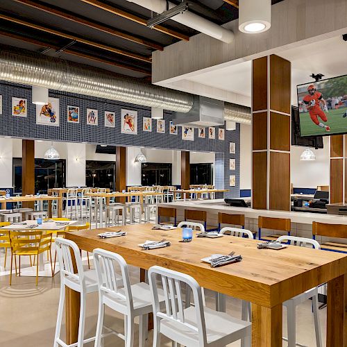 The image shows a modern sports bar with wooden tables, white chairs, a TV showing a football game, and numerous framed pictures on the walls.