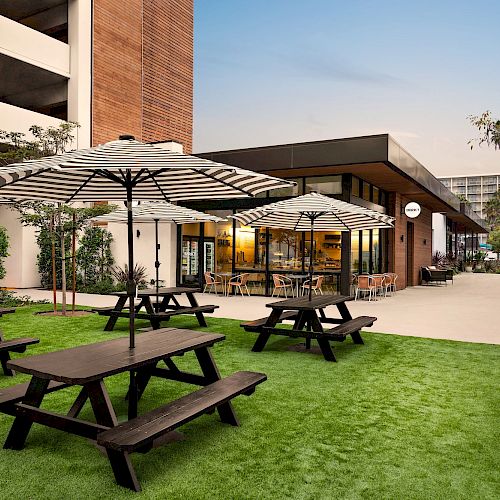An outdoor seating area with picnic tables, striped umbrellas, and artificial grass near a modern building in an urban setting.