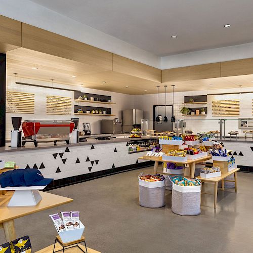 A modern coffee shop or cafe interior with various items and snacks displayed on tables, a counter with equipment, and shelves in the background.