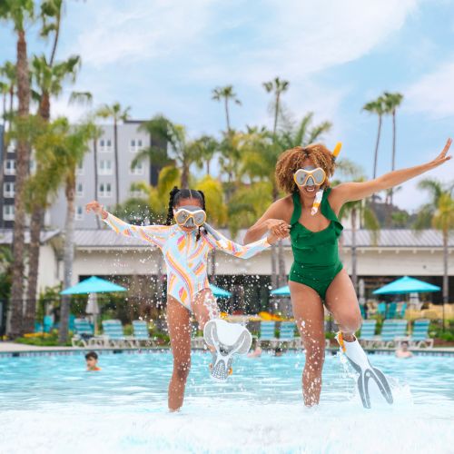 A woman and child, wearing snorkel masks and swimsuits, joyfully jump into a pool together, creating splashes, with palm trees and buildings in the background.