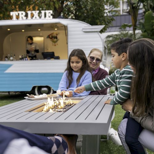 A family roasts marshmallows at a fire pit table in an outdoor setting. A food truck labeled "TRIXIE" is in the background.