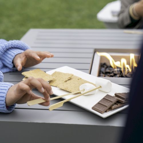 Two people are preparing s'mores with graham crackers, marshmallows, and chocolate next to a small outdoor fire pit.