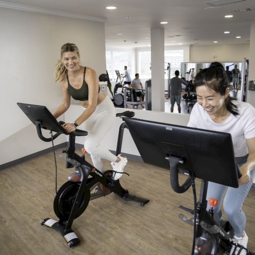 Two women are cycling on stationary bikes in a gym while looking at screens, with other gym-goers in the background.