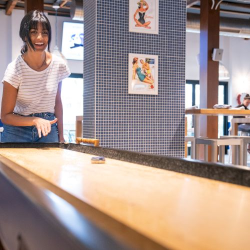 A person is playing shuffleboard in a casual setting with posters on the wall and other people in the background, enjoying their time.