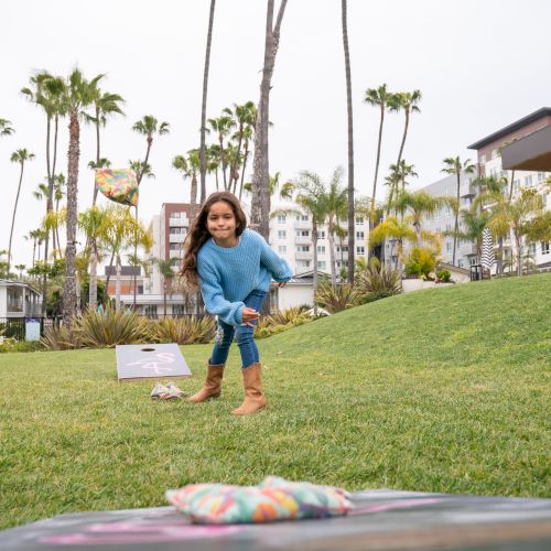 A child plays a game of cornhole in a grassy park area filled with palm trees and buildings in the background, wearing a blue sweater and boots.