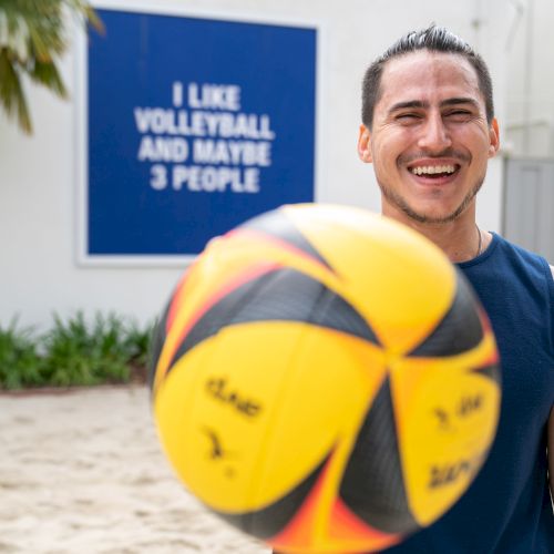 A person is smiling with a volleyball in the foreground. A sign in the background reads, "I like volleyball and maybe 3 people."
