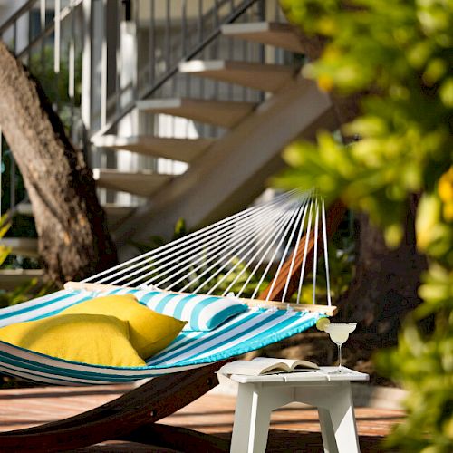 A striped hammock with yellow and green pillows, side table with a drink, and a spiral staircase in the background amidst greenery.