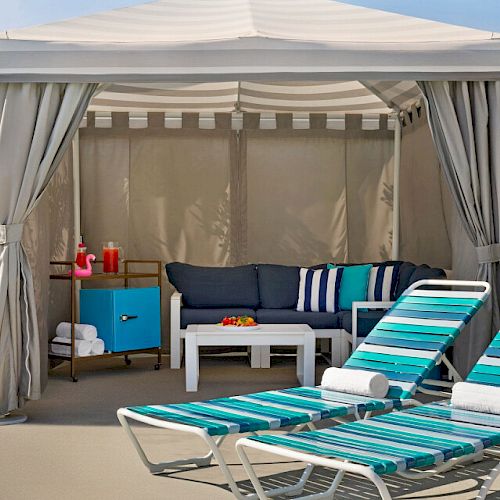 A cozy outdoor cabana with a couch, table, and two lounge chairs featuring blue-striped cushions, surrounded by greenery.