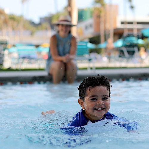 A child is happily swimming in a pool, with an adult in a hat sitting poolside in the background.