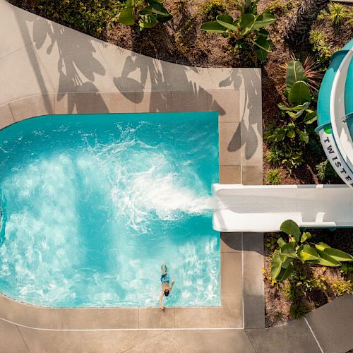 This image shows an aerial view of a swimming pool with a person swimming and a water slide. The area is surrounded by greenery and plants.