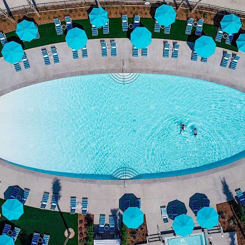 An aerial view of an oval-shaped swimming pool surrounded by numerous lounge chairs and blue umbrellas, with two people swimming in the pool.