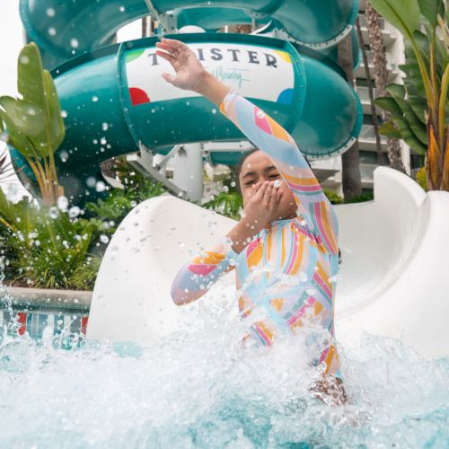 A person is splashing out of a water slide in a colorful outfit at a water park, covering their face with their hand.