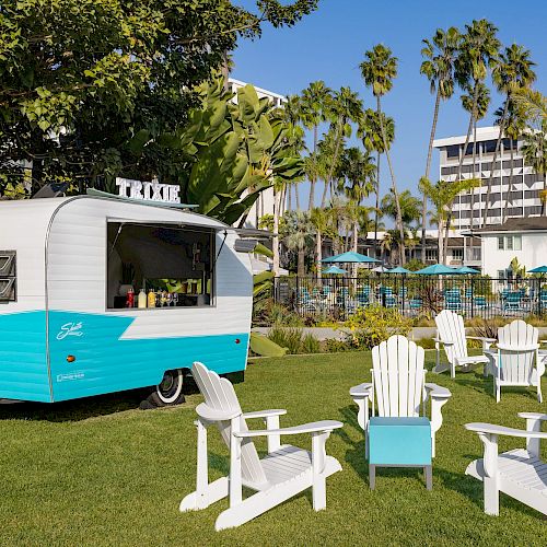 A vintage turquoise and white camper is parked on a grassy area surrounded by white Adirondack chairs, with a pool and buildings nearby.