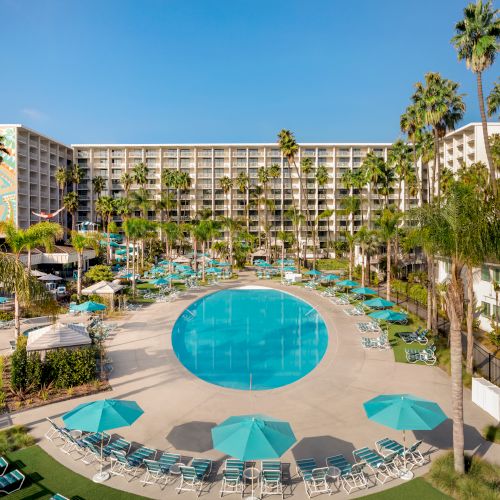 This image displays a large hotel with multiple floors, featuring a circular pool, numerous lounge chairs, umbrellas, and palm trees surrounding it.