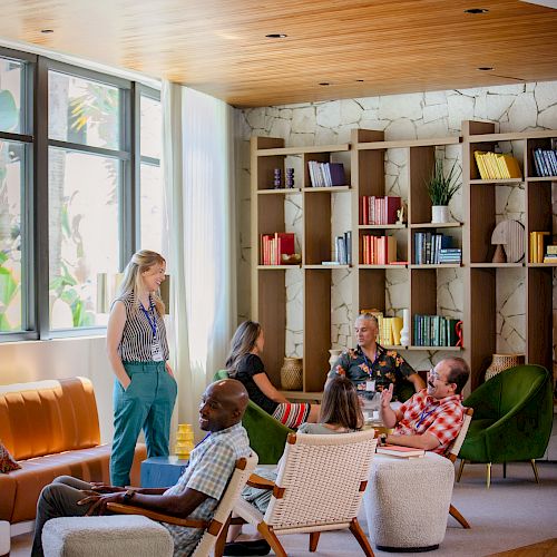 People are sitting and standing in a modern lounge area with bookshelves, relaxed seating, and a wooden ceiling, engaged in conversation.