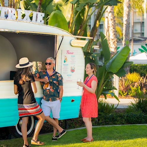 Three people are standing outside a colorful food truck named "Trixie," set in a sunny, tropical environment with palm trees and lush greenery.