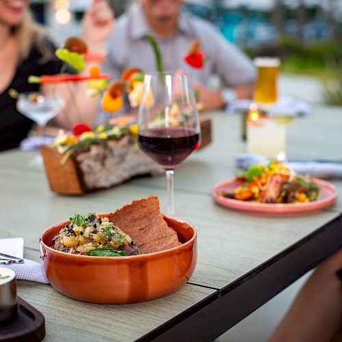 People are enjoying a meal with various dishes and drinks on an outdoor table. A glass of red wine is prominently featured in the foreground.