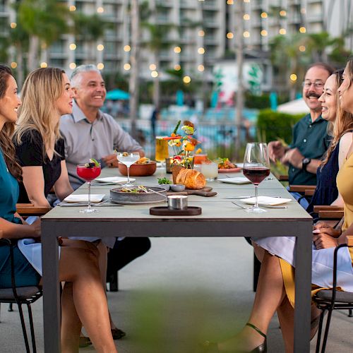Six people are seated around an outdoor dining table, enjoying food and drinks, with a background of lights and greenery, ending the sentence.