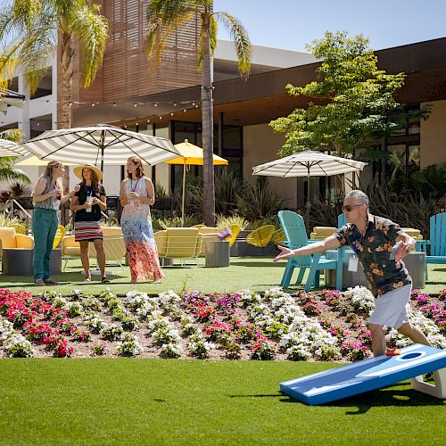 A man is playing cornhole on a sunny day, while three people watch and smile near a garden, surrounded by outdoor furniture and umbrellas.