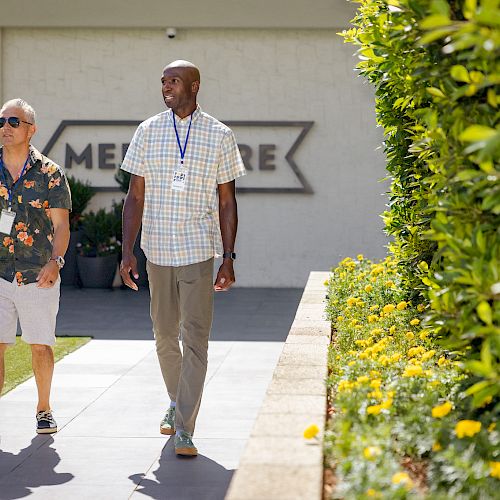 Two people are walking outdoors on a pathway beside green bushes and flowers. A sign behind them reads "Medical Care."