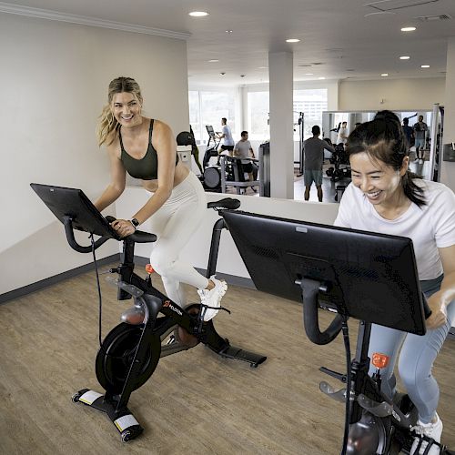 Two women are smiling and riding stationary bikes in a gym with other people using various fitness equipment in the background.