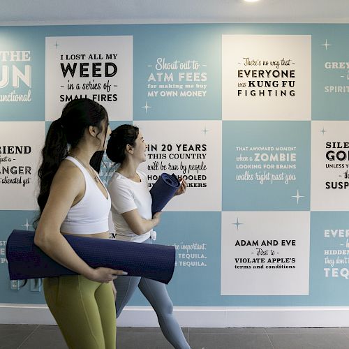 Two people carrying yoga mats walk past a wall decorated with humorous and motivational quotes in a gym or wellness center.