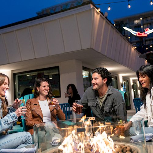 Four people sitting around a fire pit, enjoying drinks and each other's company in an outdoor setting with string lights hanging above them.