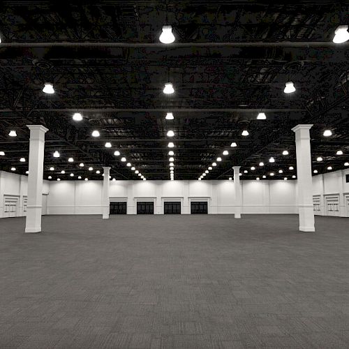 The image shows a large, empty convention center or warehouse space with numerous ceiling lights and support columns.