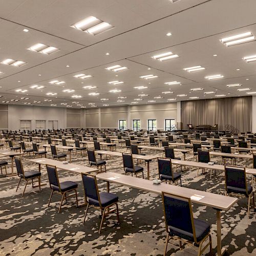 The image shows a large, empty conference room set up with numerous rows of tables and chairs facing a stage, suggesting preparation for a seminar or event.