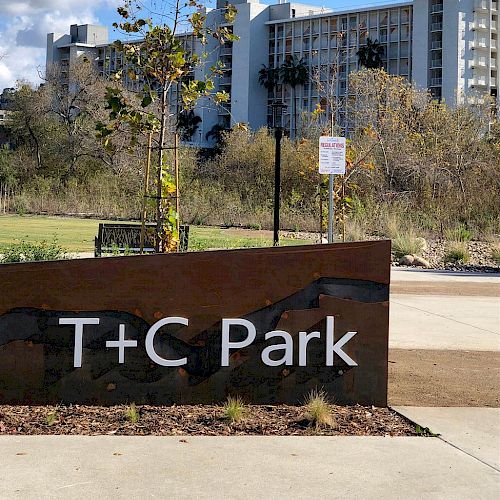 The image shows an entrance sign for "T+C Park" with a cityscape in the background and some greenery around the area, including trees and a bench.