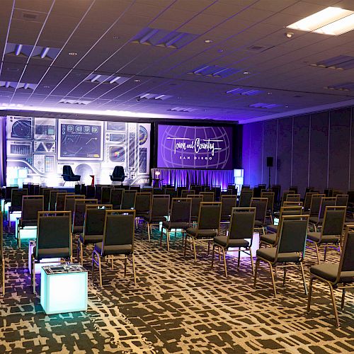 A conference room with rows of chairs facing a stage with two armchairs, multiple screens, and purple lighting.