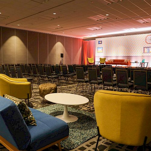 The image shows a conference room setup with rows of chairs facing a stage. The stage has decor, chairs, and a projection screen, ending the sentence.